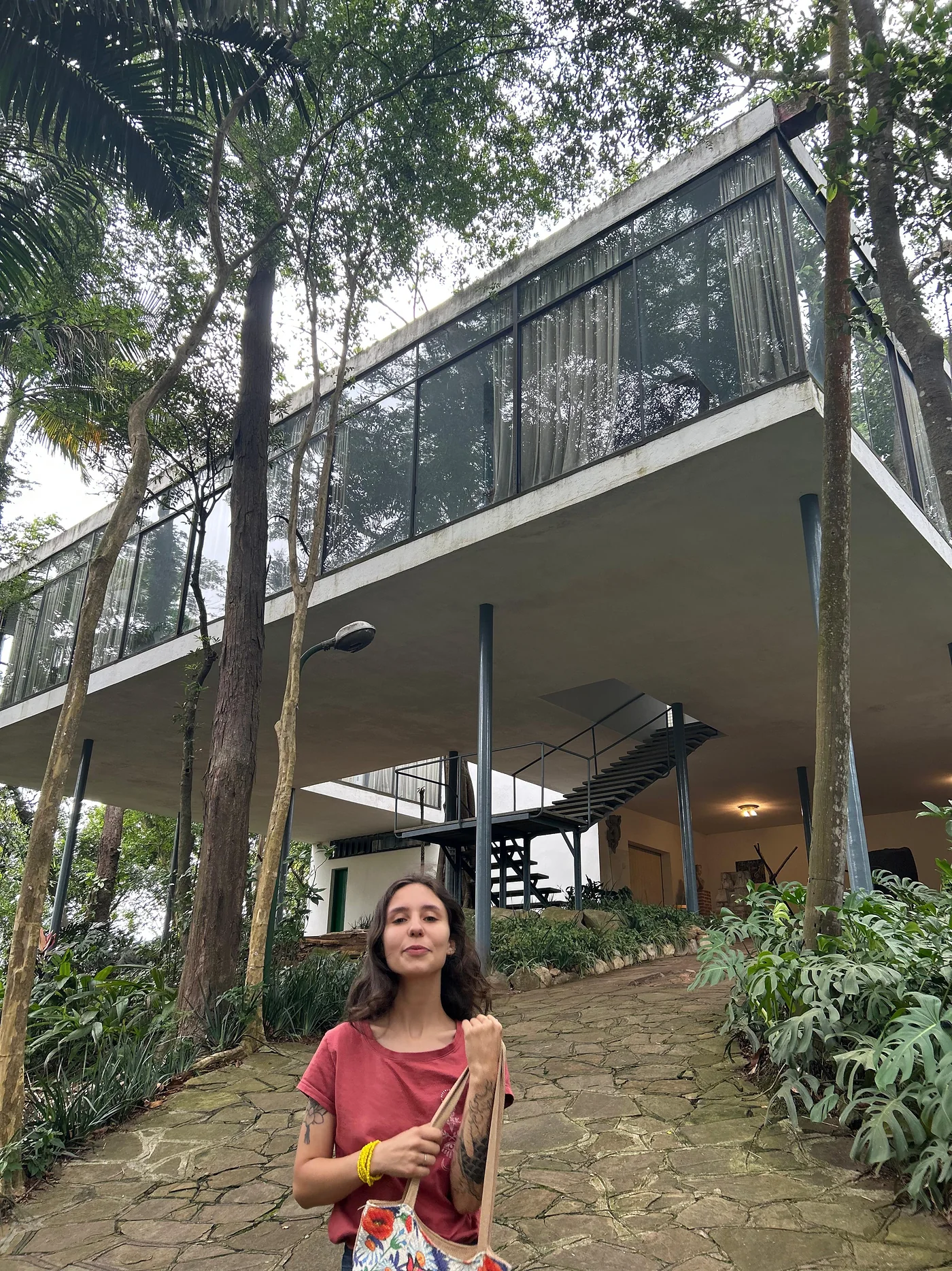 Karen de camiseta vermelha sob uma casa suspensa sobre pilotis, entre árvores.