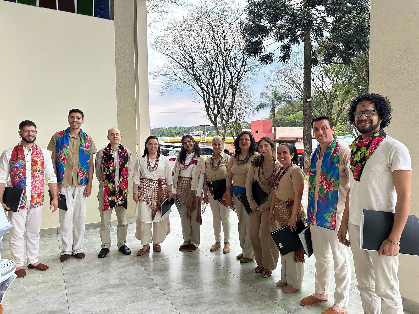 O coro reunido num pátio coberto, de roupas claras, lenços floridos e colares; cada cantor segura sua pasta de partituras.