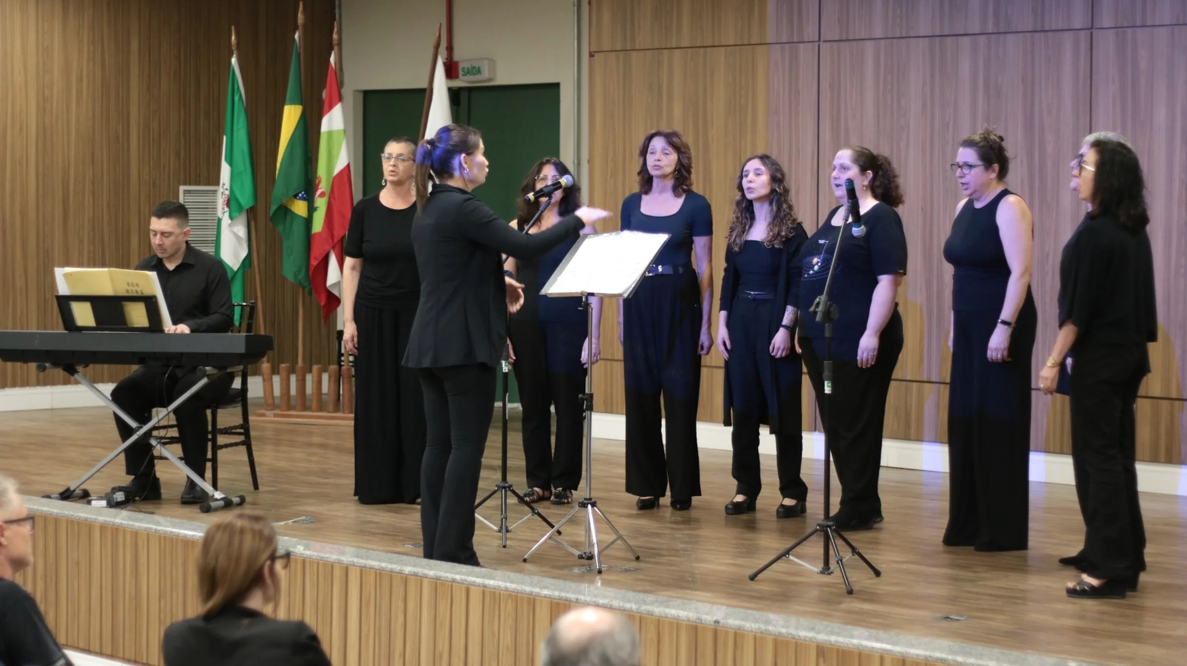 Coral feminino em auditório, com regente e pianista.