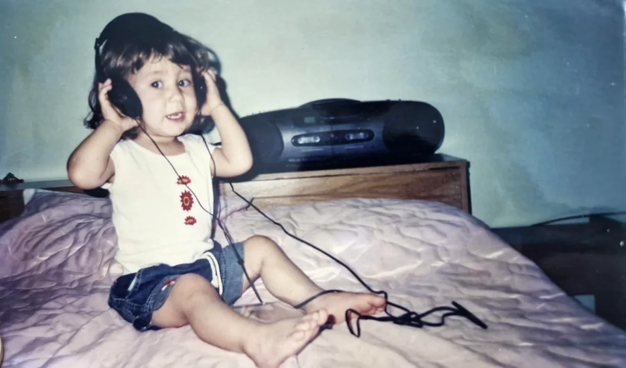 Fotografia antiga: Karen bebê, sentada numa cama, ouve música com fones de ouvido ligados a um aparelho de som.