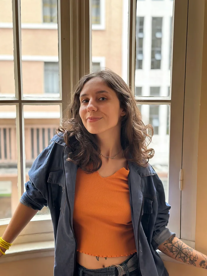 Karen de blusa laranja e jaqueta jeans, diante de uma janela.