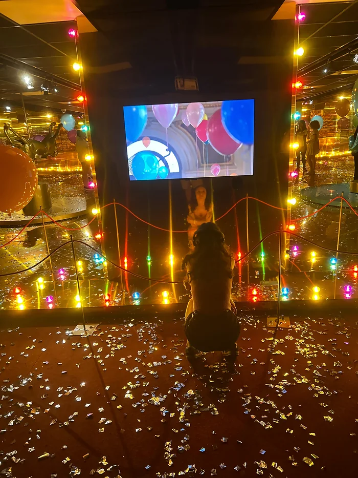 Karen de costas, num salão iluminado por lâmpadas coloridas.