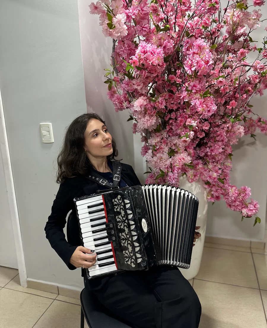 Karen toca acordeom ao lado de flores cor-de-rosa.