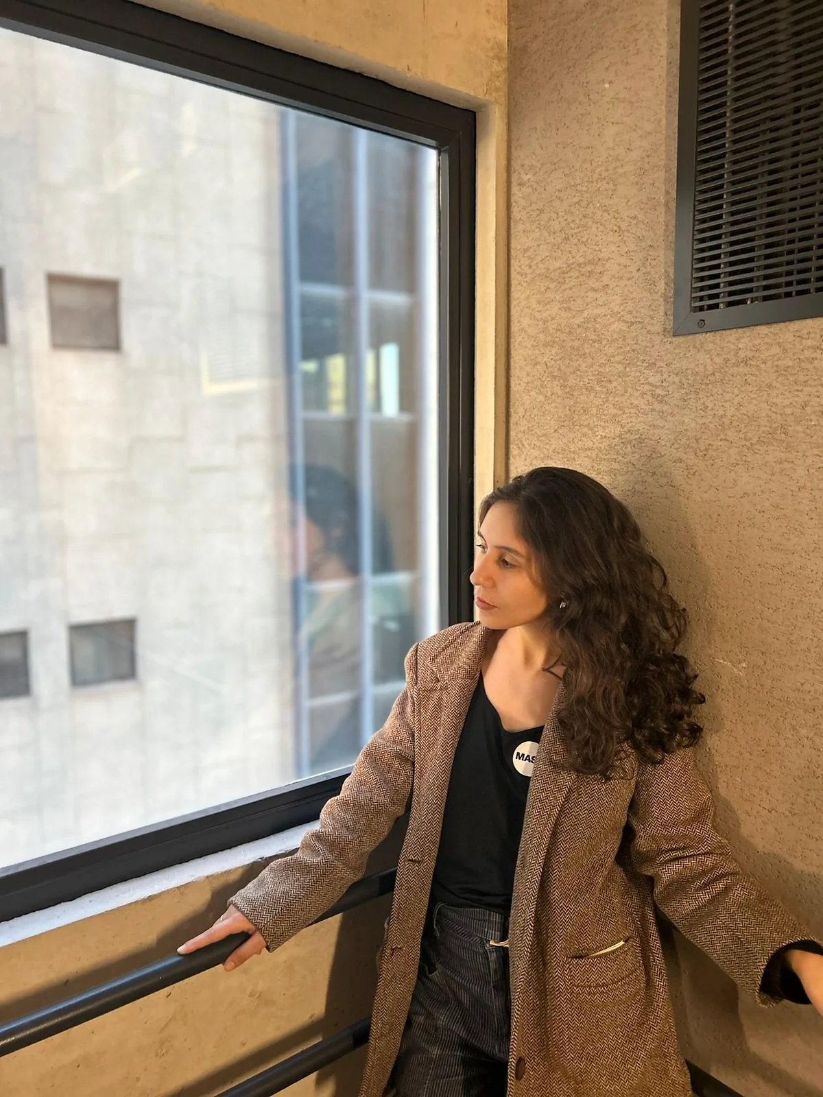 Karen de blazer, encostada junto a uma janela alta, olha para fora.